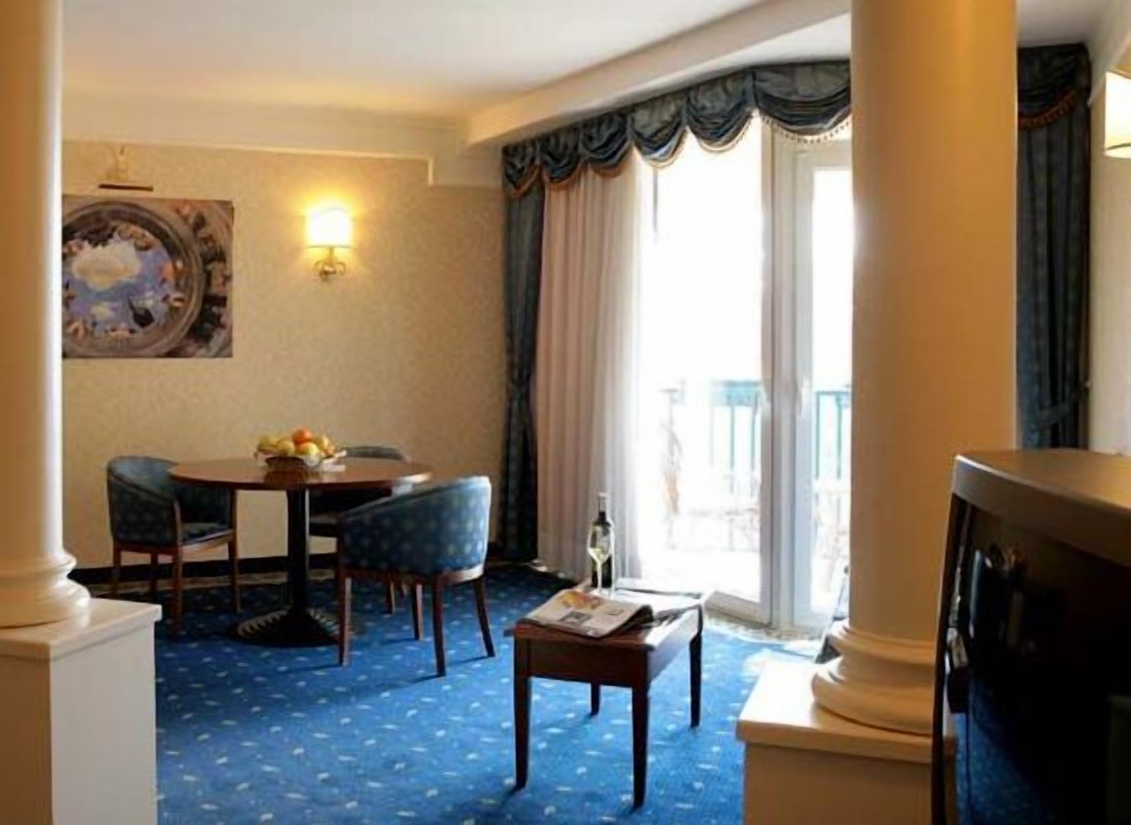 A hotel room with a dining area, a television, a window with curtains, and a painting on the wall.