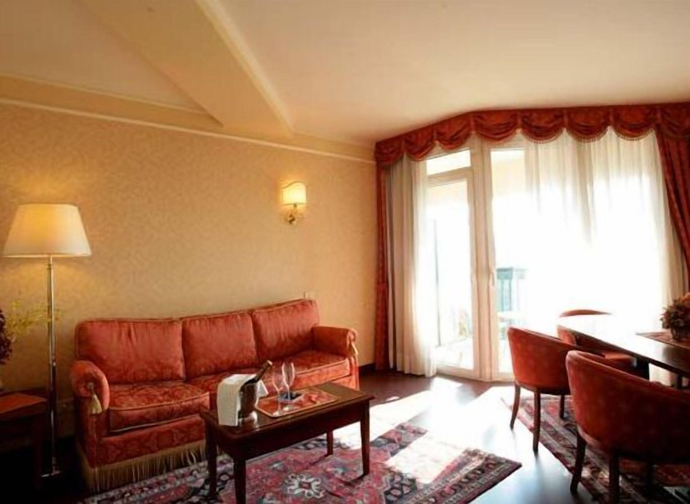 Un salon avec un canapé rouge, une table basse et une lampe. Il y a une fenêtre avec des rideaux et une vue sur l’extérieur.