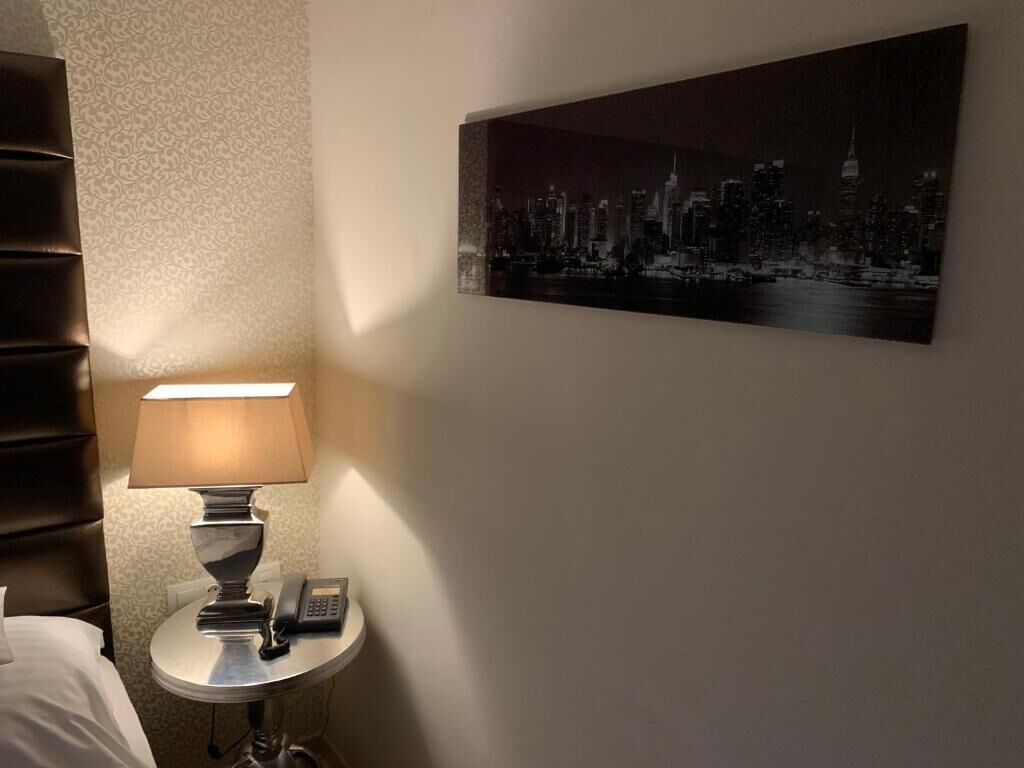 Habitación de hotel con una cama, una mesita de noche con lámpara, un teléfono y un cuadro de paisaje urbano en la pared.