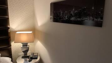 Habitación de hotel con una cama, una mesita de noche con lámpara, un teléfono y un cuadro de paisaje urbano en la pared.