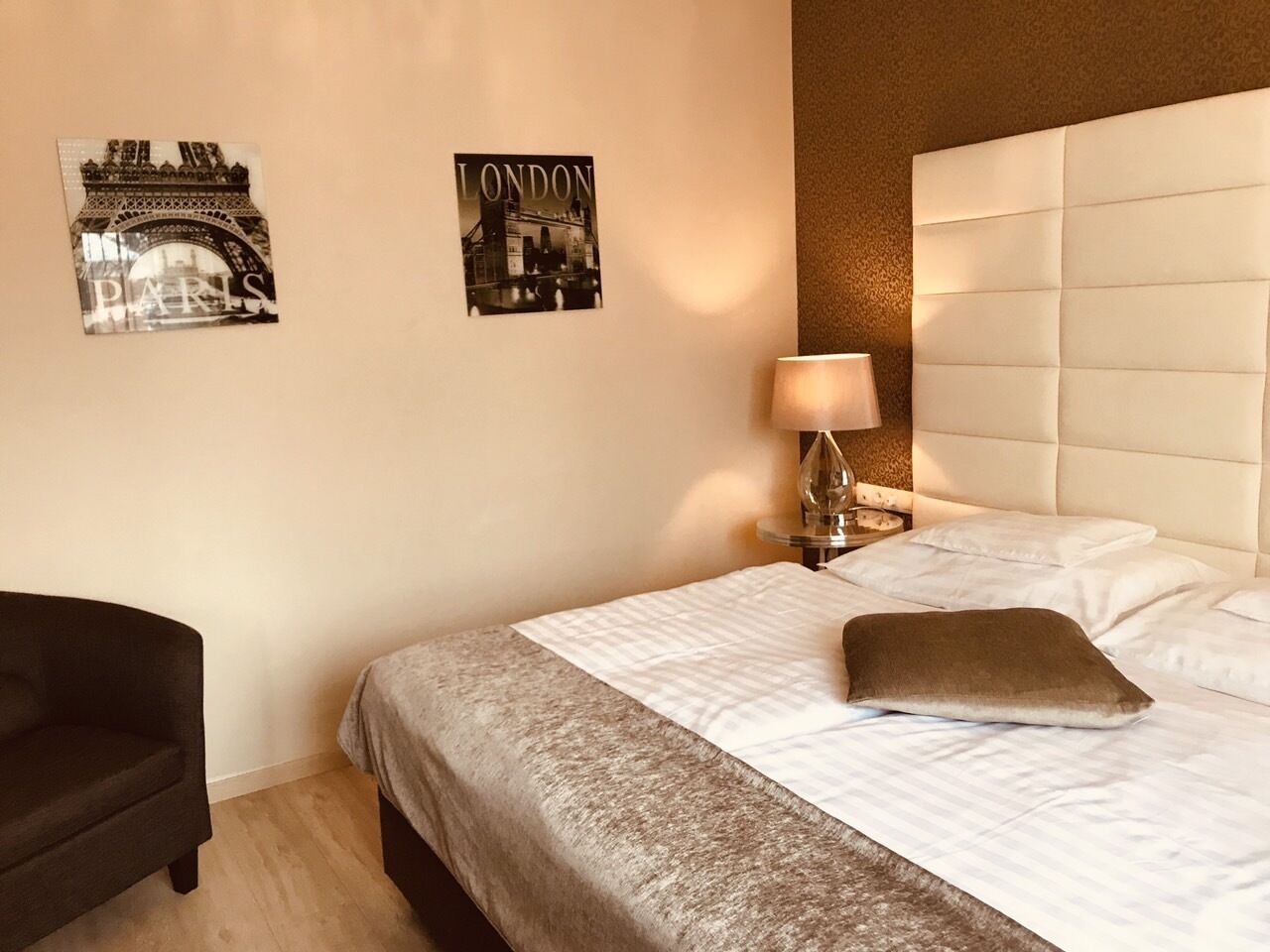 Habitación de hotel con una cama, una mesita de noche con lámpara, una silla y dos cuadros en la pared de París y Londres.