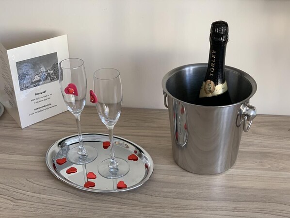 Een fles champagne in een metalen emmer, twee glazen op een schaal met rode hartdecoraties, en een kaart met een foto en tekst.