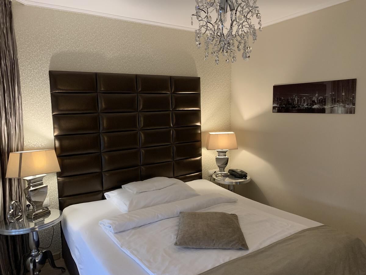 Habitación de hotel con una cama grande, dos mesitas de noche, una araña y un cuadro de paisaje urbano en la pared.