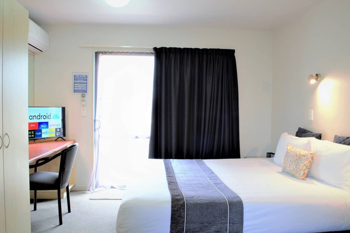 A hotel room with a bed, a desk with a computer, a chair, a window with curtains, and a wall-mounted light.