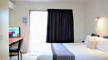 A hotel room with a bed, a desk with a computer, a chair, a window with curtains, and a wall-mounted light.
