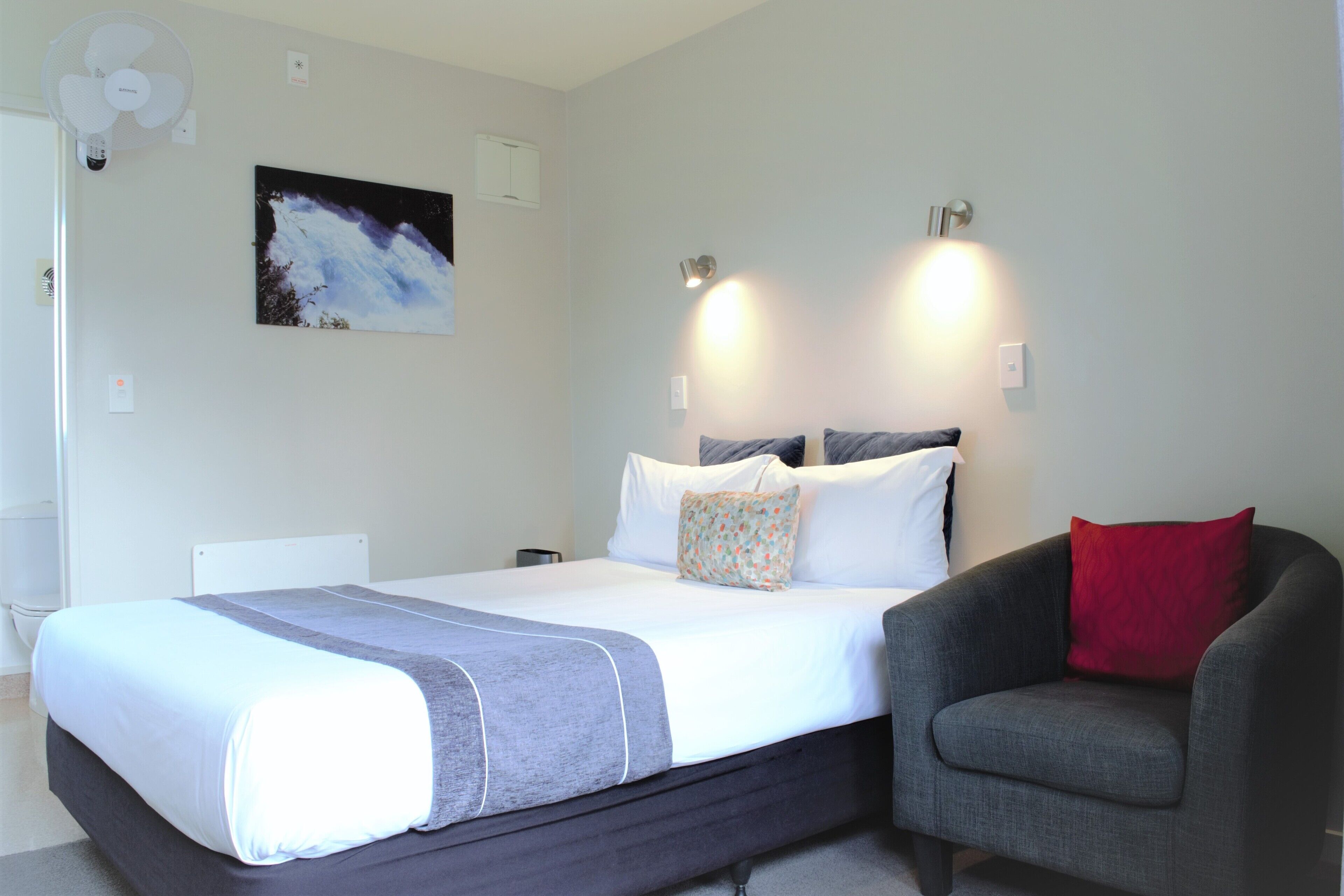 Ein Schlafzimmer mit einem Bett, einem Sessel, einem Wandventilator, einem Gemälde und zwei Wandleuchten.