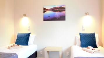 A hotel room with two beds, each with a pillow and a towel. There is a small table between the beds, and a painting of a landscape on the wall.