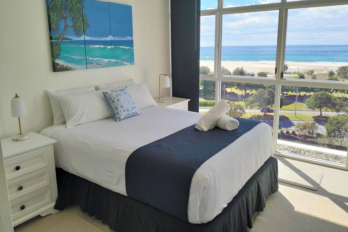 A bedroom with a large window, a bed with white and navy bedding, a painting of a tropical beach, and a nightstand with a lamp.