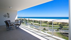 A balcony with a table and chairs, overlooking a coastal road and beach. - Kirra Surf Apartments (Coolangatta)