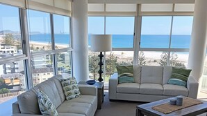 A modern living room with a large window overlooking a beach, two sofas, a coffee table, and a lamp. - Kirra Surf Apartments (Coolangatta)