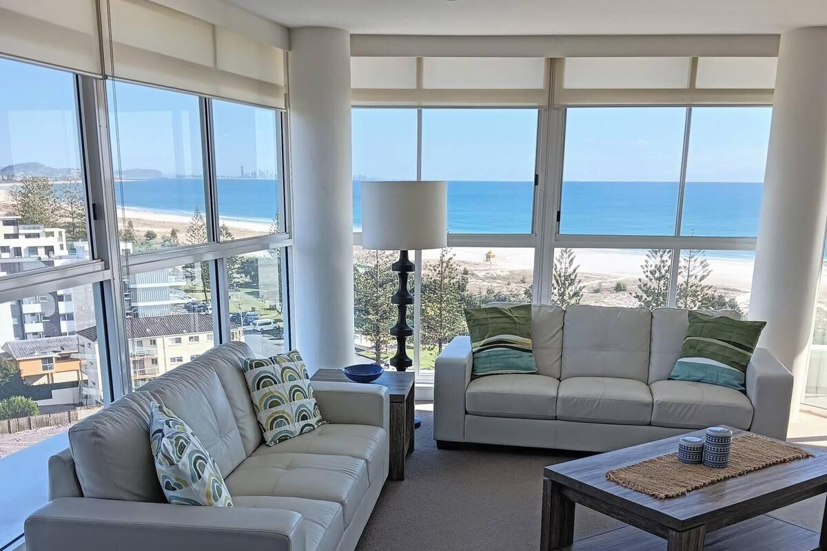 A modern living room with a large window overlooking a beach, two sofas, a coffee table, and a lamp.