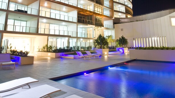 A modern hotel with a curved facade, multiple balconies, and a swimming pool area with lounge chairs. - Kirra Surf Apartments (Coolangatta)