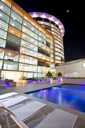 A modern hotel with a curved facade, multiple balconies, and a swimming pool area with lounge chairs. - Kirra Surf Apartments (Coolangatta)