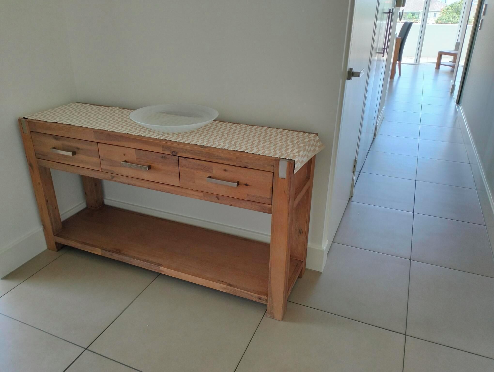 Una scrivania in legno con piano intrecciato e un oggetto circolare bianco sopra, situata in un corridoio con pavimento piastrellato.