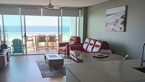 A modern living room with a view of the ocean, a red sofa, a dining area, and a kitchen with a dishwasher. - Kirra Surf Apartments (Coolangatta)