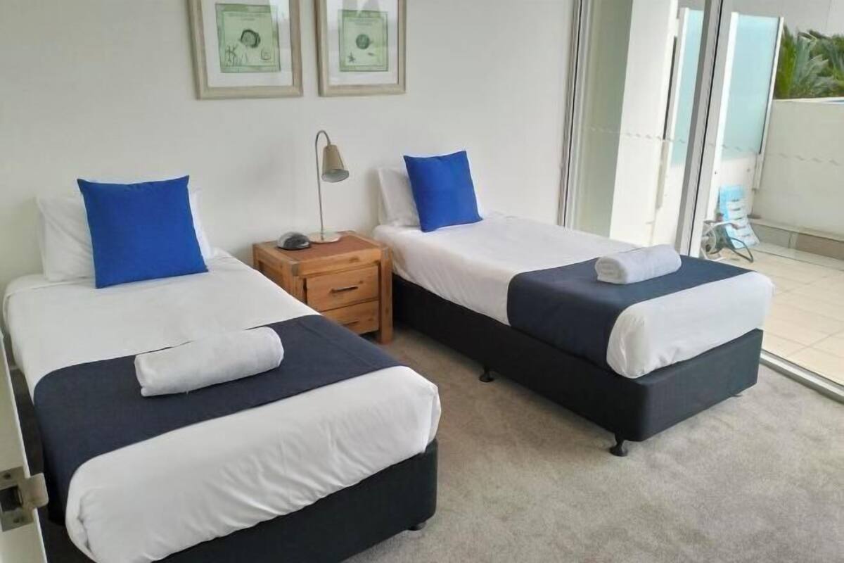 A hotel room with two beds, a nightstand, a lamp, and a sliding glass door leading to a balcony.