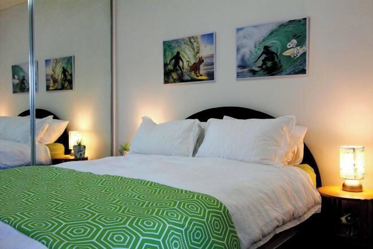 A neatly made bed with a green patterned bedspread, two bedside tables with lamps, and three wall-mounted artworks depicting surfing scenes.