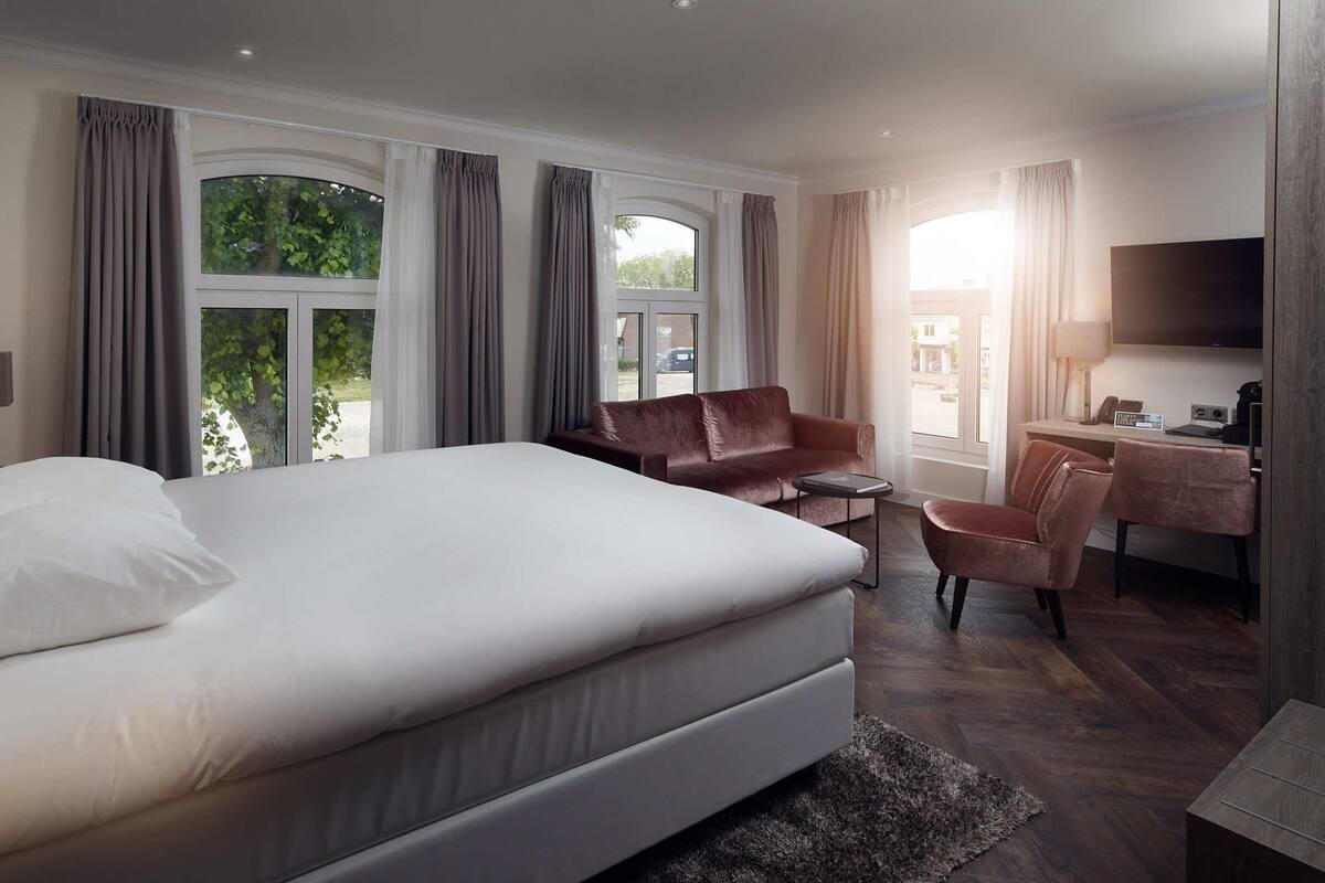 A modern hotel room with a large bed, a leather sofa, a desk with a chair, and a TV.