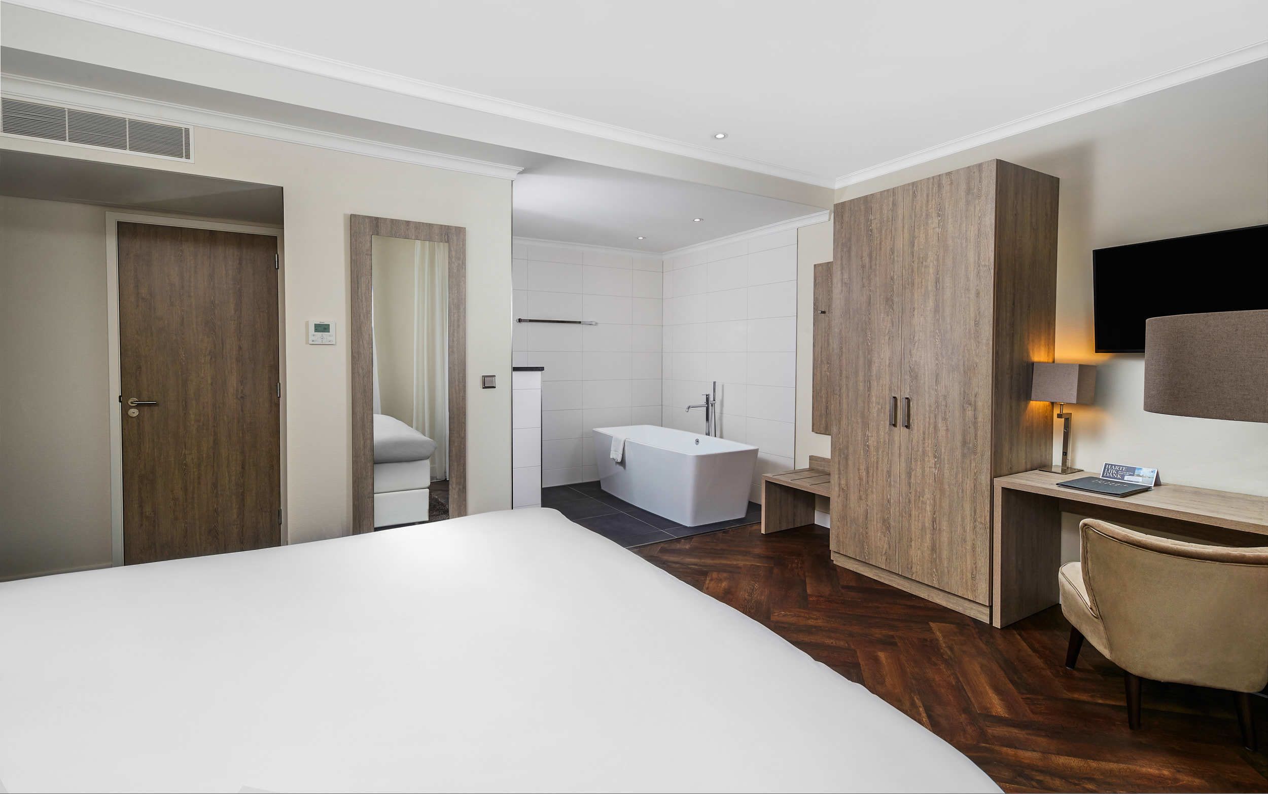 Una habitación de hotel moderna con una cama grande, un armario de madera, un escritorio con silla, un televisor de pantalla plana y un baño separado con bañera.