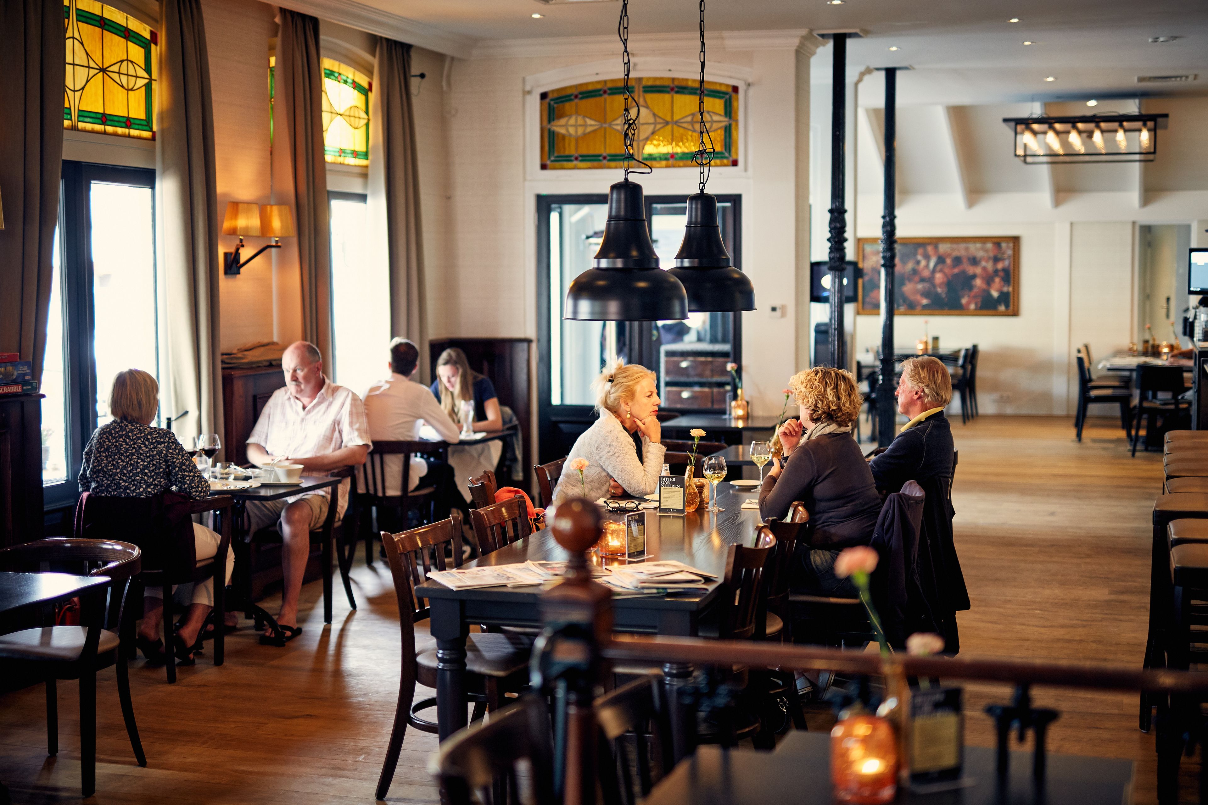Um restaurante com mesas e cadeiras de madeira, janelas de vitral e luminárias pendentes.