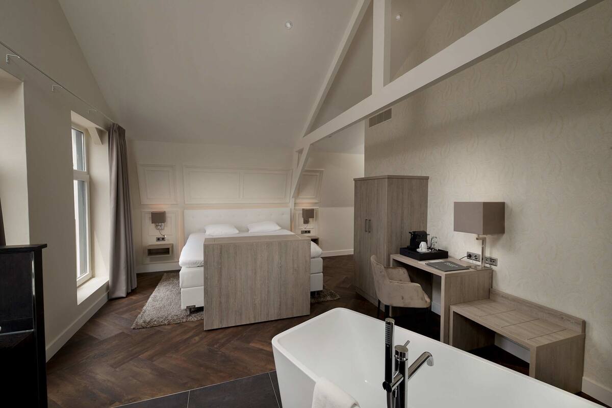 A modern hotel room with a large bed, a desk, a chair, a bathtub, and a wardrobe.