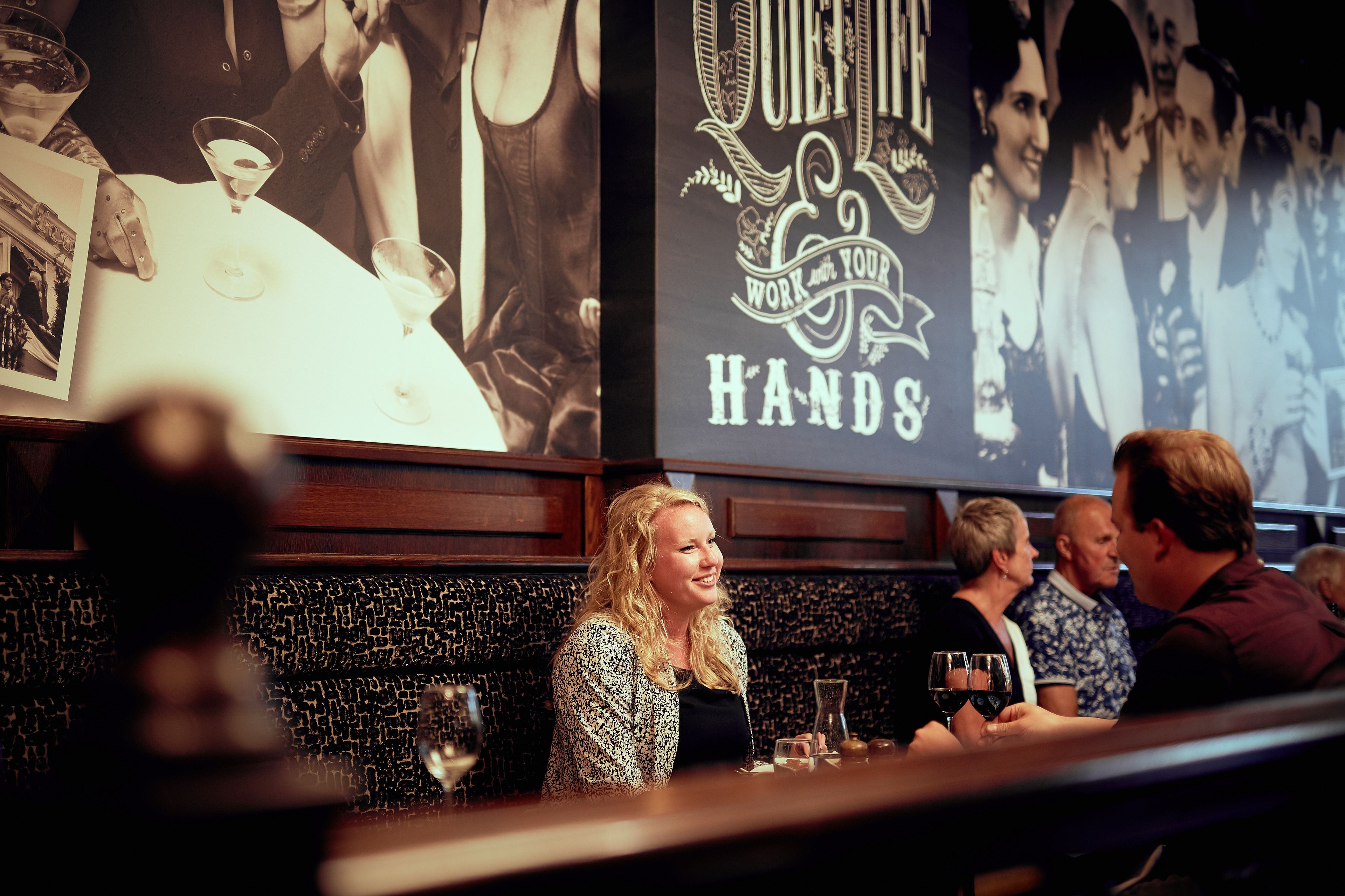 A bar with a black and white patterned wall, a poster with the text "QUILT YOUR HANDS," and a group of people seated at the bar.