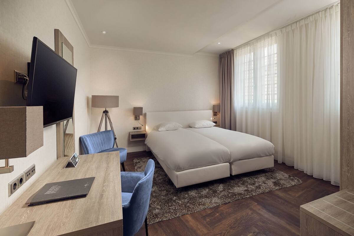 A hotel room with a bed, a desk with a laptop, a chair, a TV, and a nightstand with lamps.