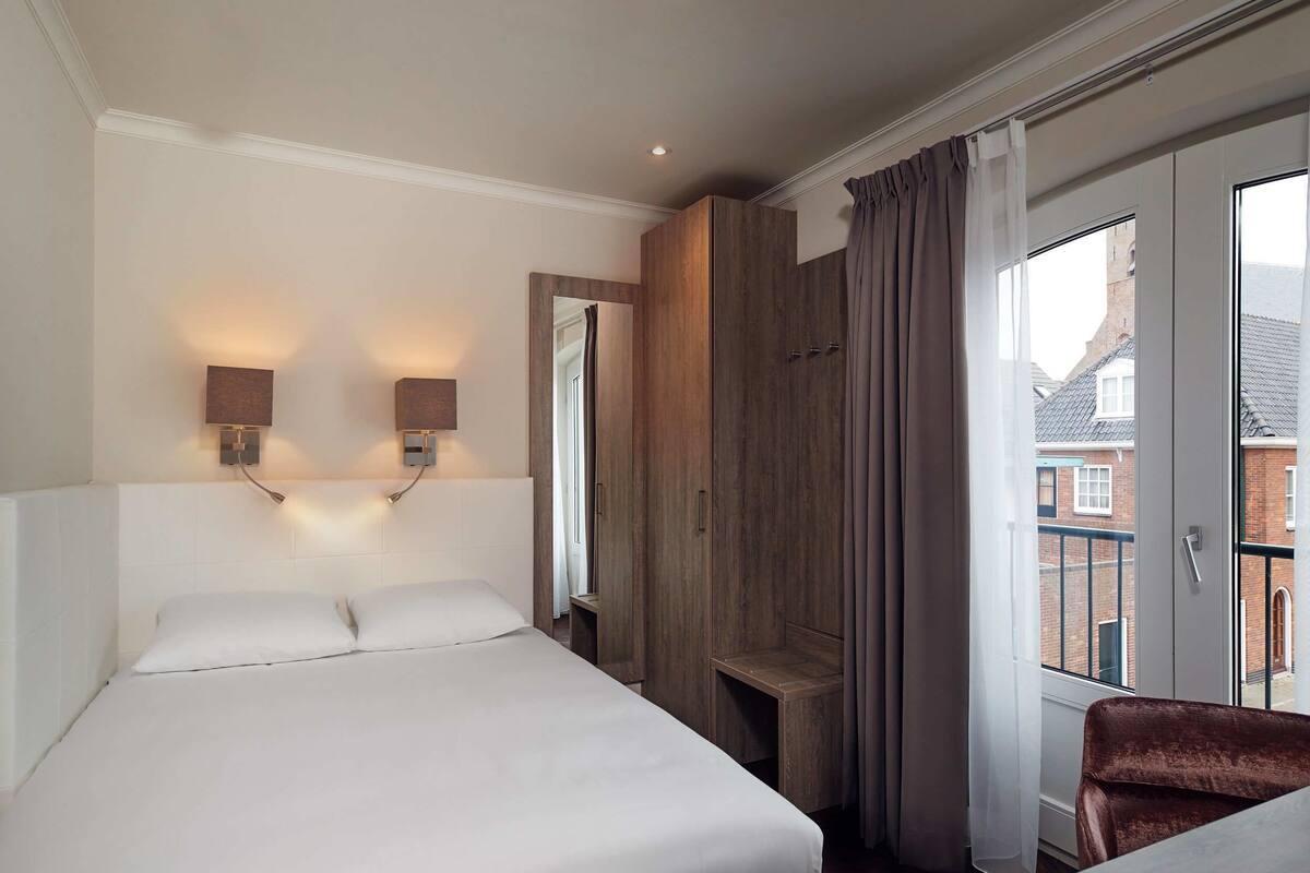 A hotel room with a large bed, a wooden wardrobe, a mirror, and a window with curtains.