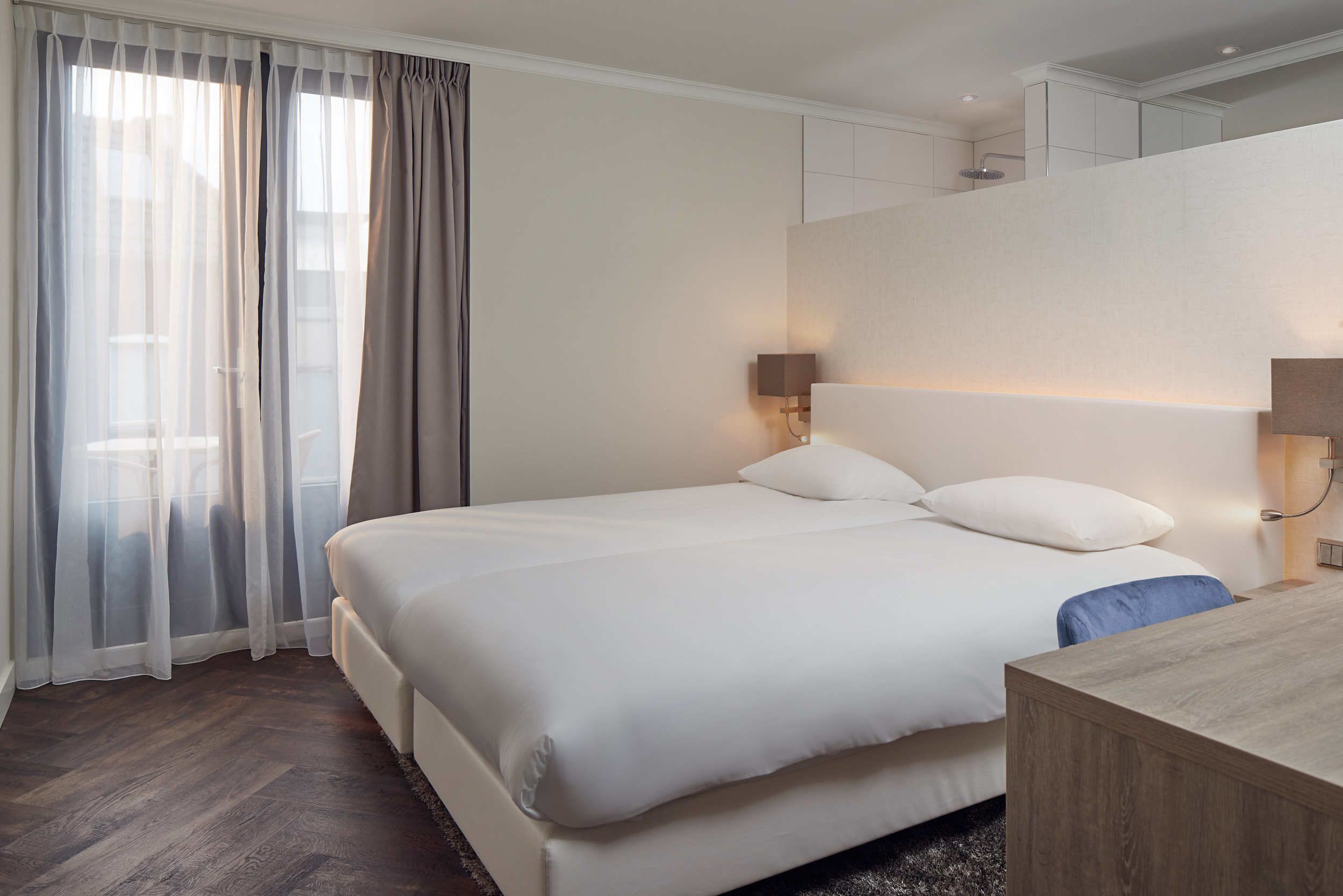 A modern hotel room with a large bed, a wooden desk, and a view of the city through sheer curtains.