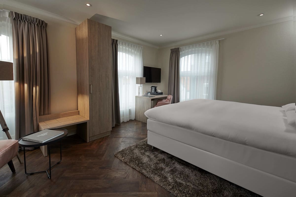 A modern hotel room with a large bed, a desk with a lamp, a TV, and a seating area with curtains.