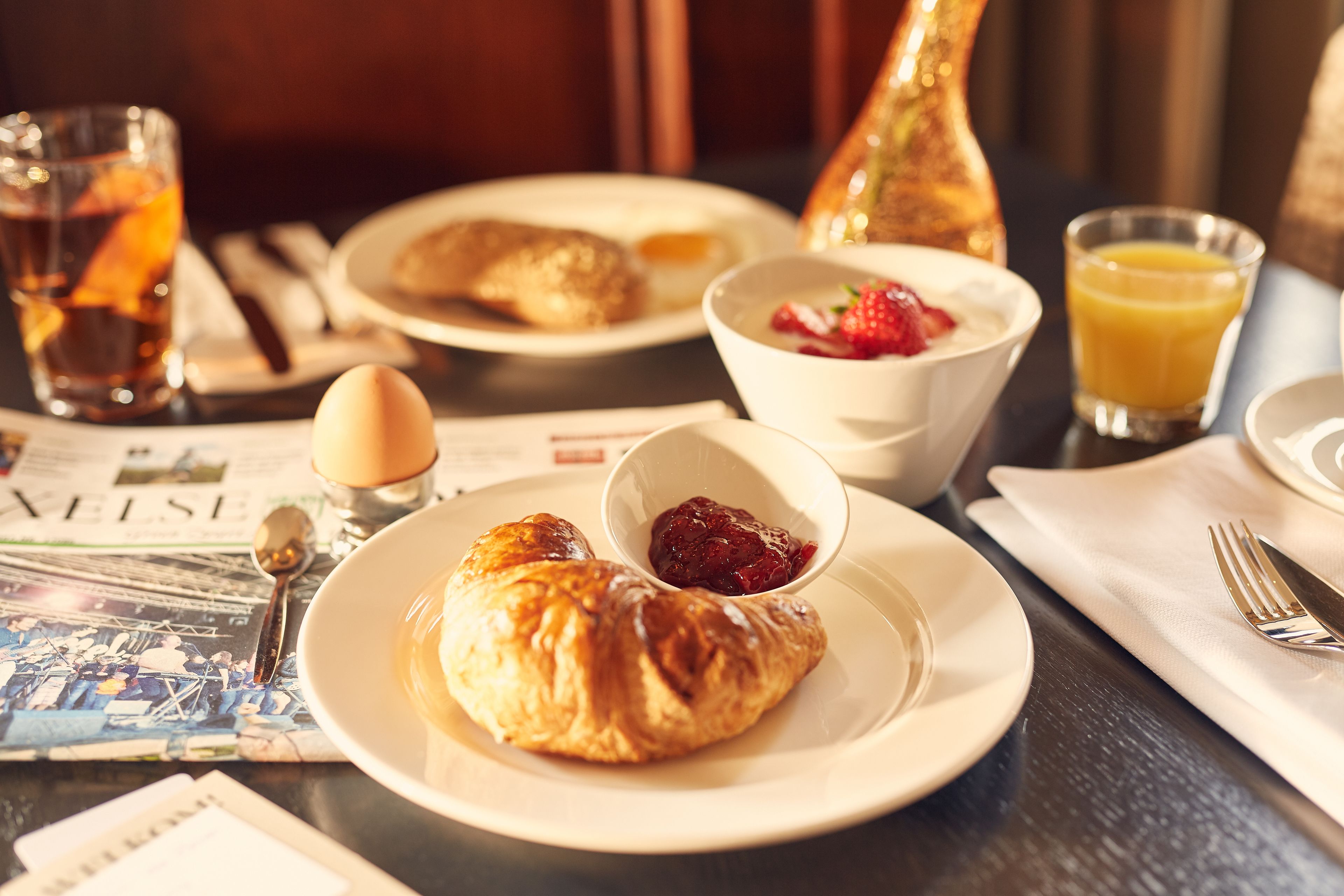 Un desayuno con croissants, un bol de compota de frutas, un vaso de jugo de naranja y un periódico.
