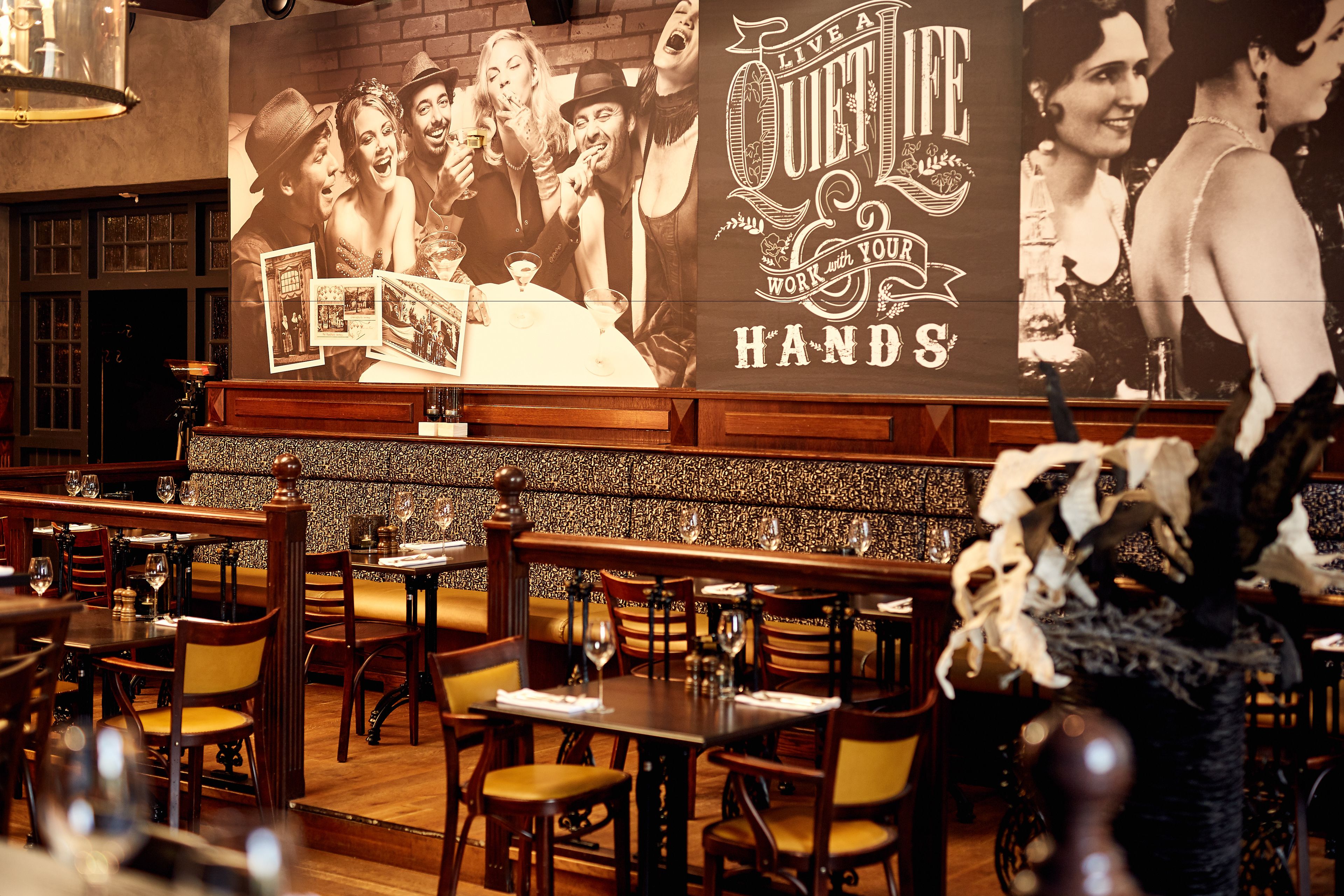 A restaurant interior with wooden tables and chairs, a brick wall, and a large poster advertising 'Live a Quiet Life'.