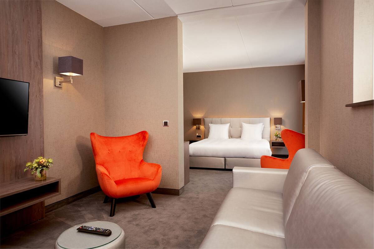 A hotel room with a modern design, featuring a large bed, a sofa, an orange armchair, a flat-screen TV, and a small table.