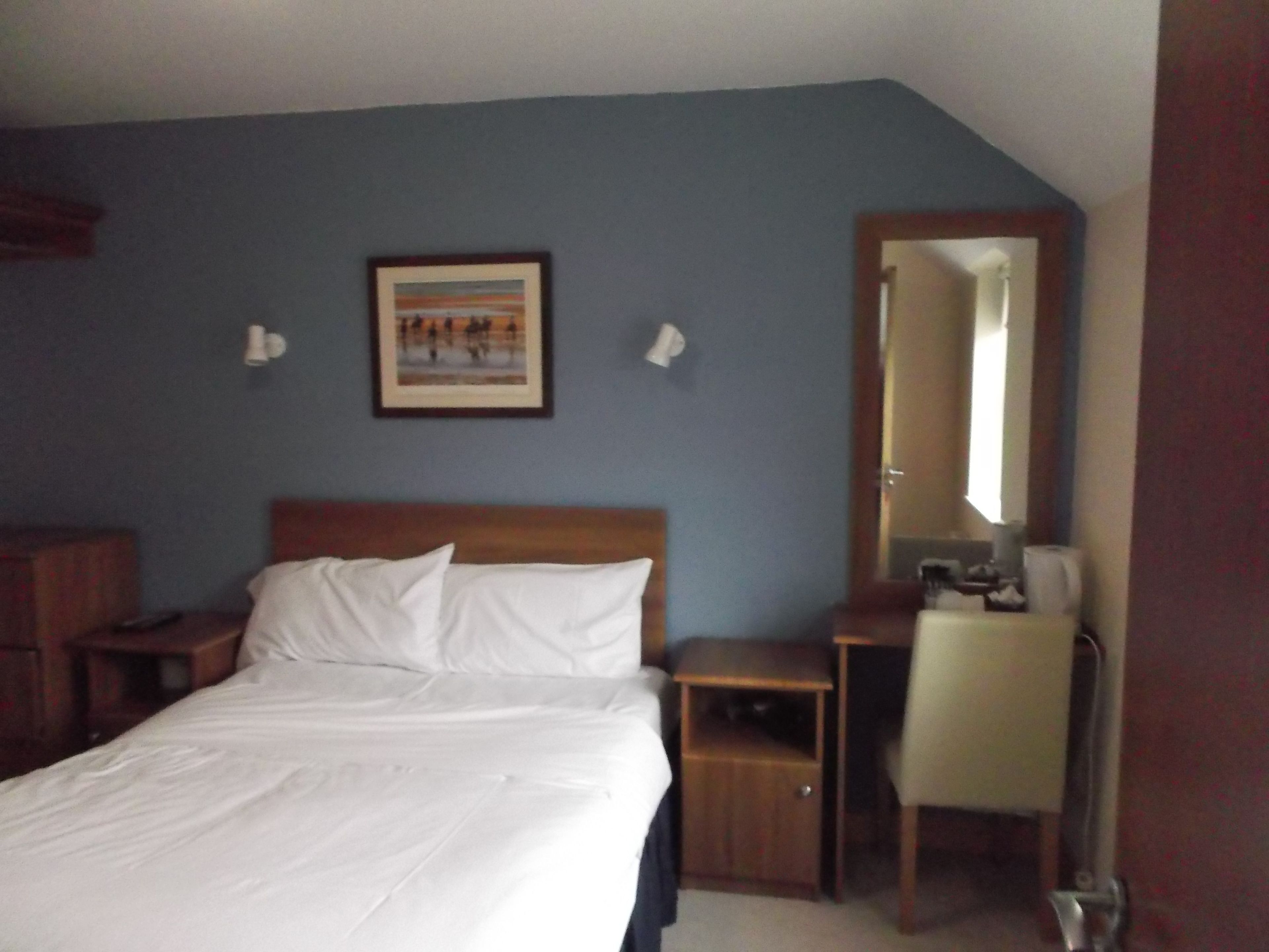 Une chambre d’hôtel comprenant un lit, un bureau, une chaise, une table de chevet, une lampe de chevet, un tableau encadré au mur et un miroir.