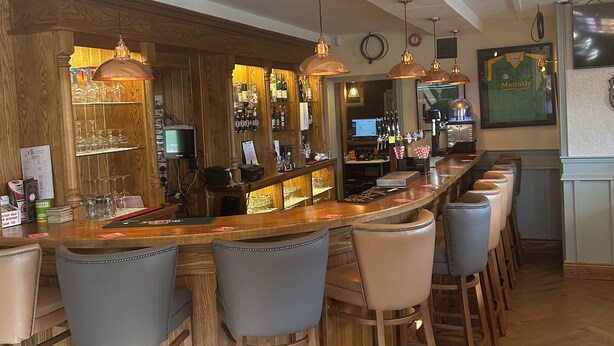 Zona de bar con taburetes de madera, mostrador con caja registradora y diferentes botellas y vasos.