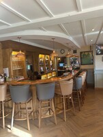 Zona de bar con taburetes de madera, mostrador con caja registradora y diferentes botellas y vasos.