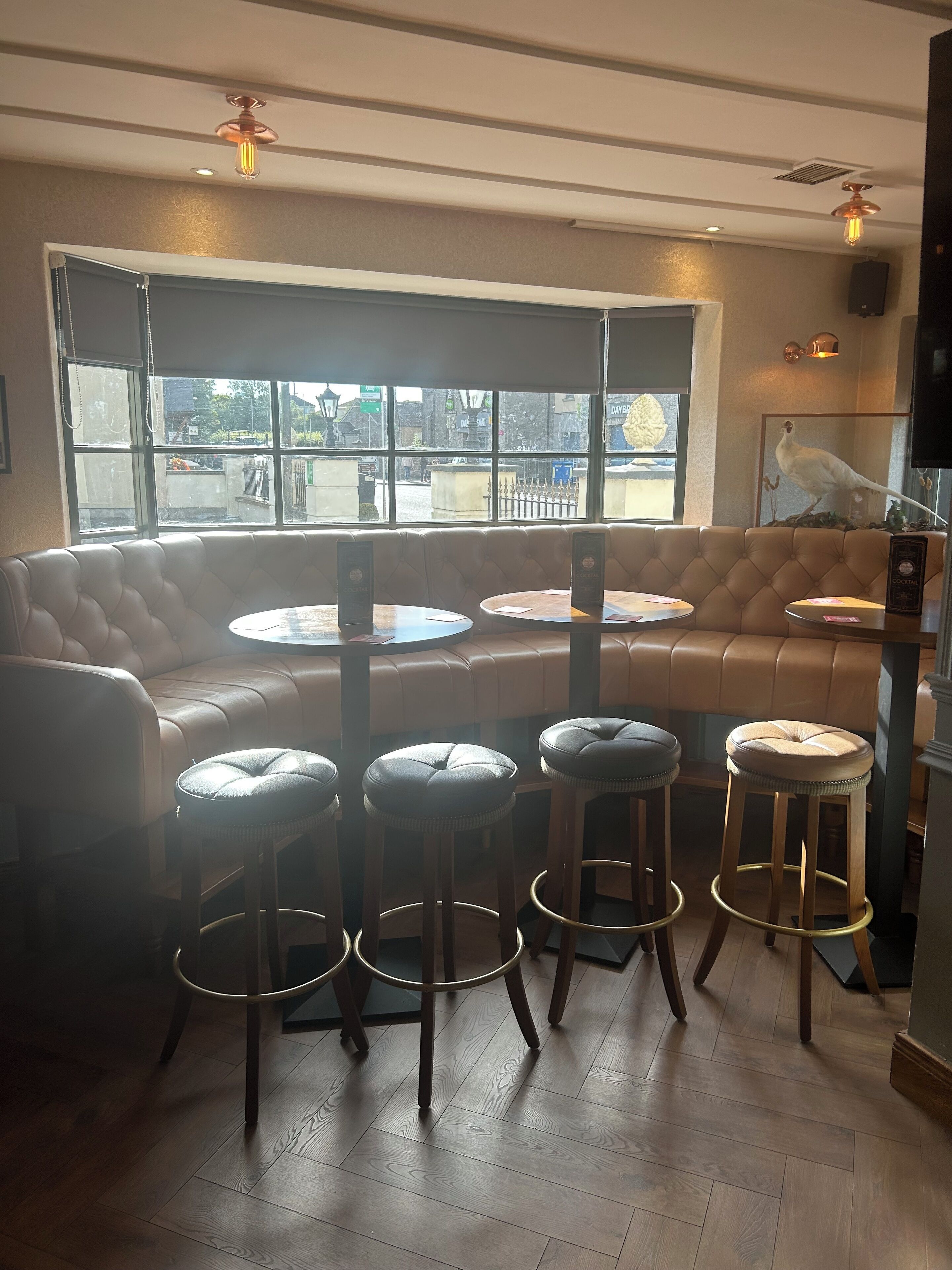A cozy seating area with a leather couch and round tables, wooden stools, and a large window offering a view of the street.