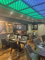 Un restaurante con suelo de madera, sillas tapizadas y mesas. Cuenta con una zona de bar con mostrador y carta. El techo presenta un diseño geométrico.