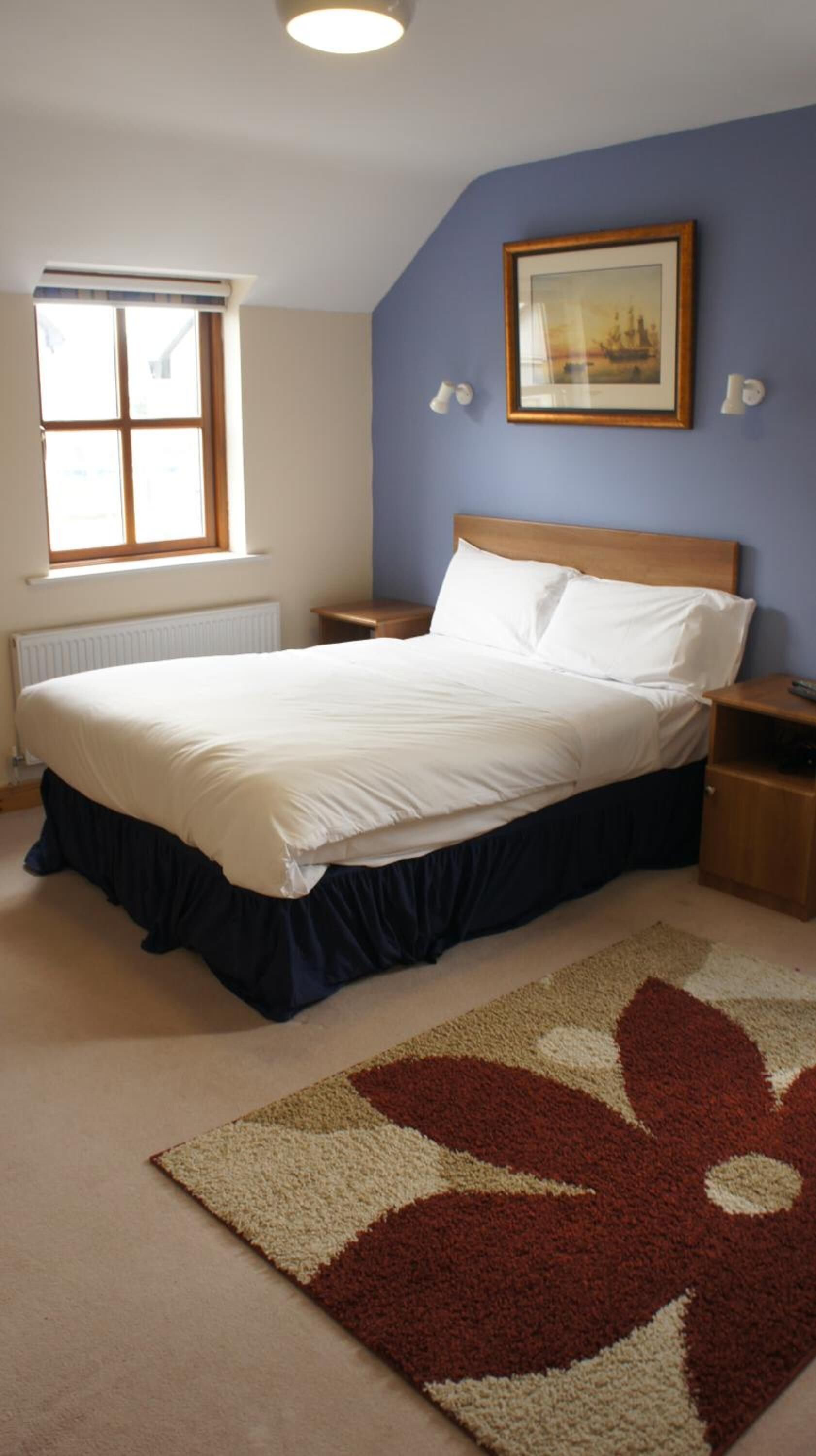 Un dormitorio con cama, mesita de noche, un cuadro enmarcado en la pared, ventana y alfombra estampada.