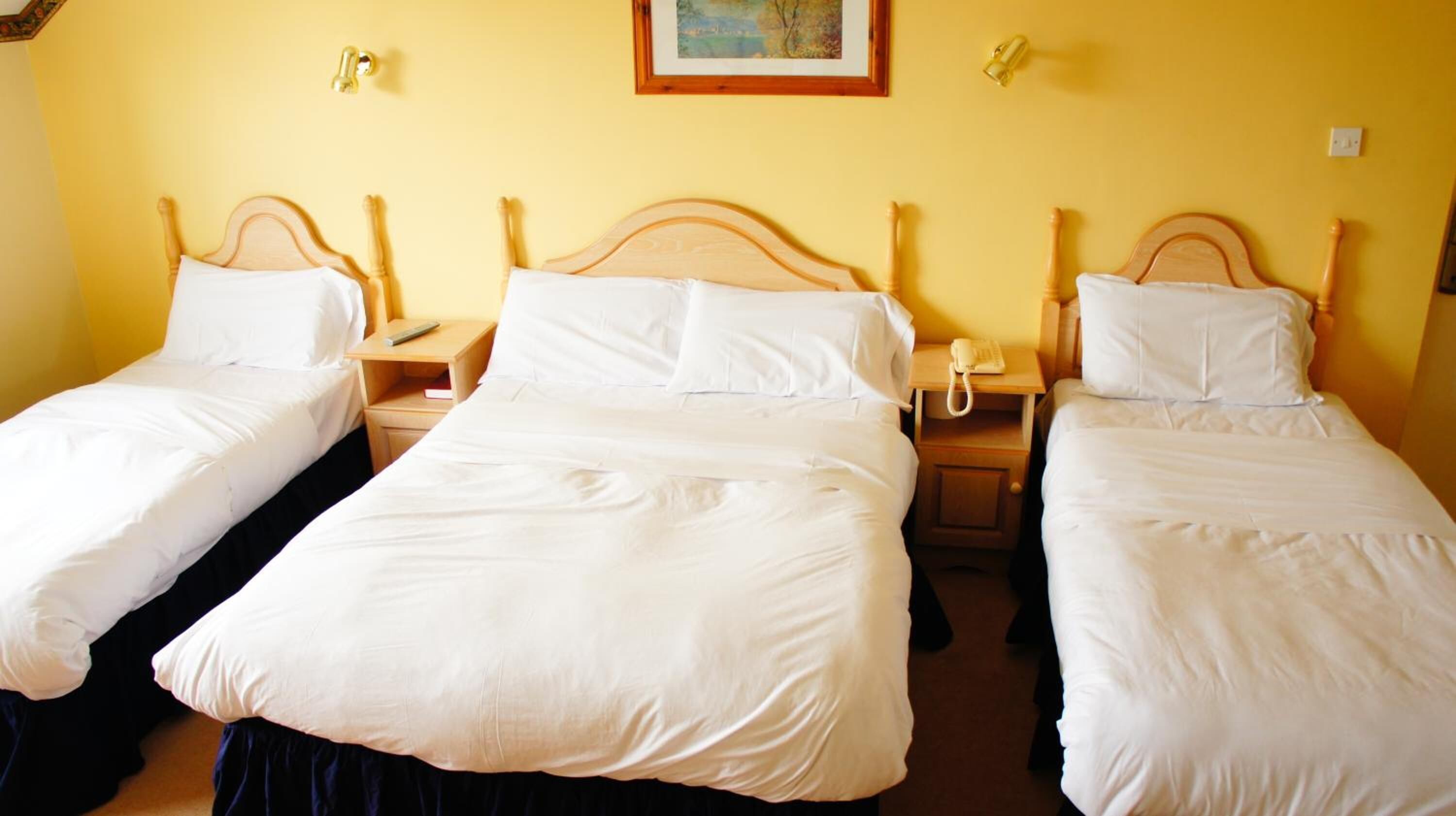 Habitación de hotel con tres camas individuales, cada una con cabecera de madera y ropa de cama blanca. Hay mesitas de noche con lámparas y teléfono.
