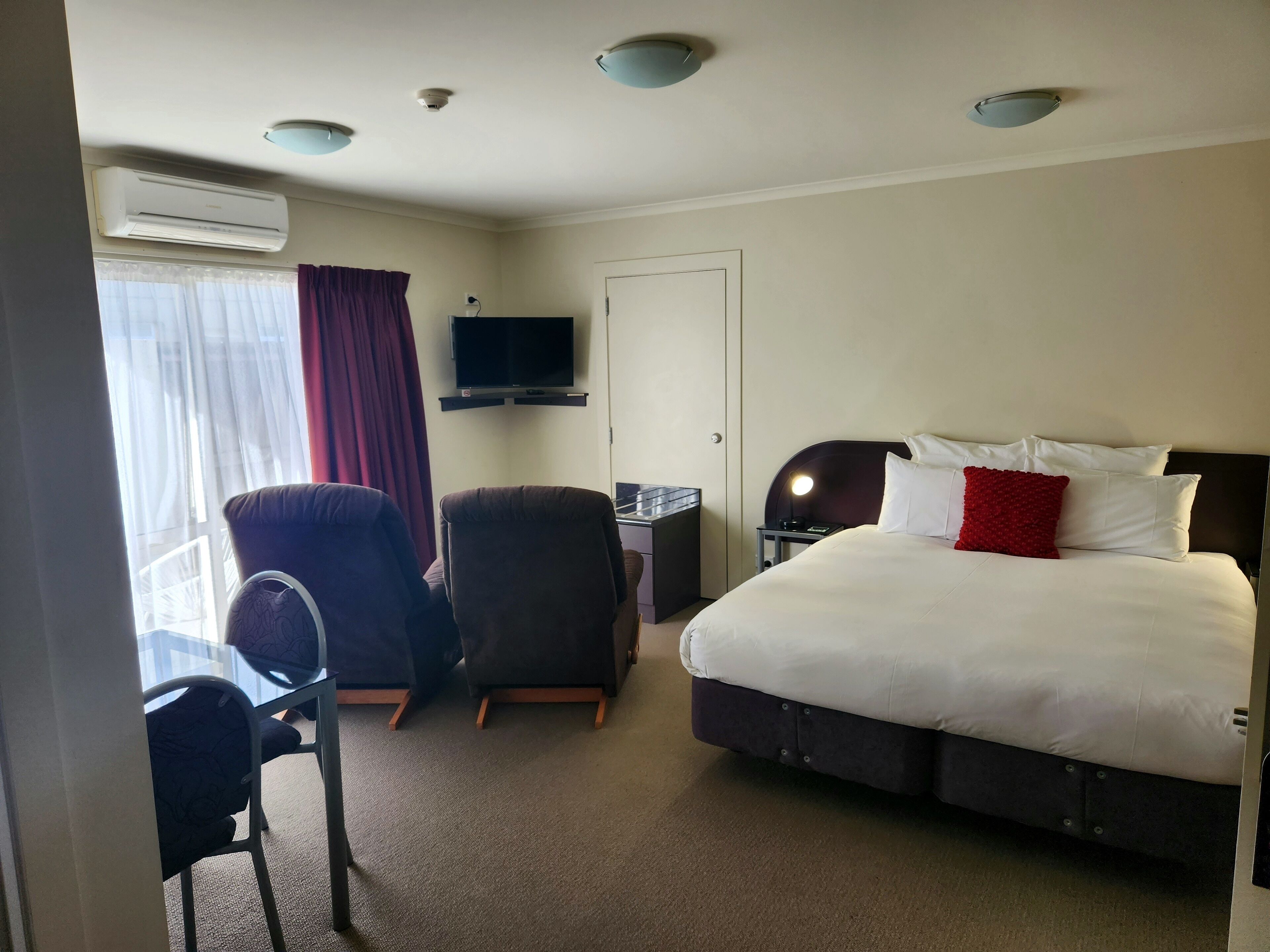 A hotel room with a bed, two chairs, a desk, a TV, and an air conditioning unit.