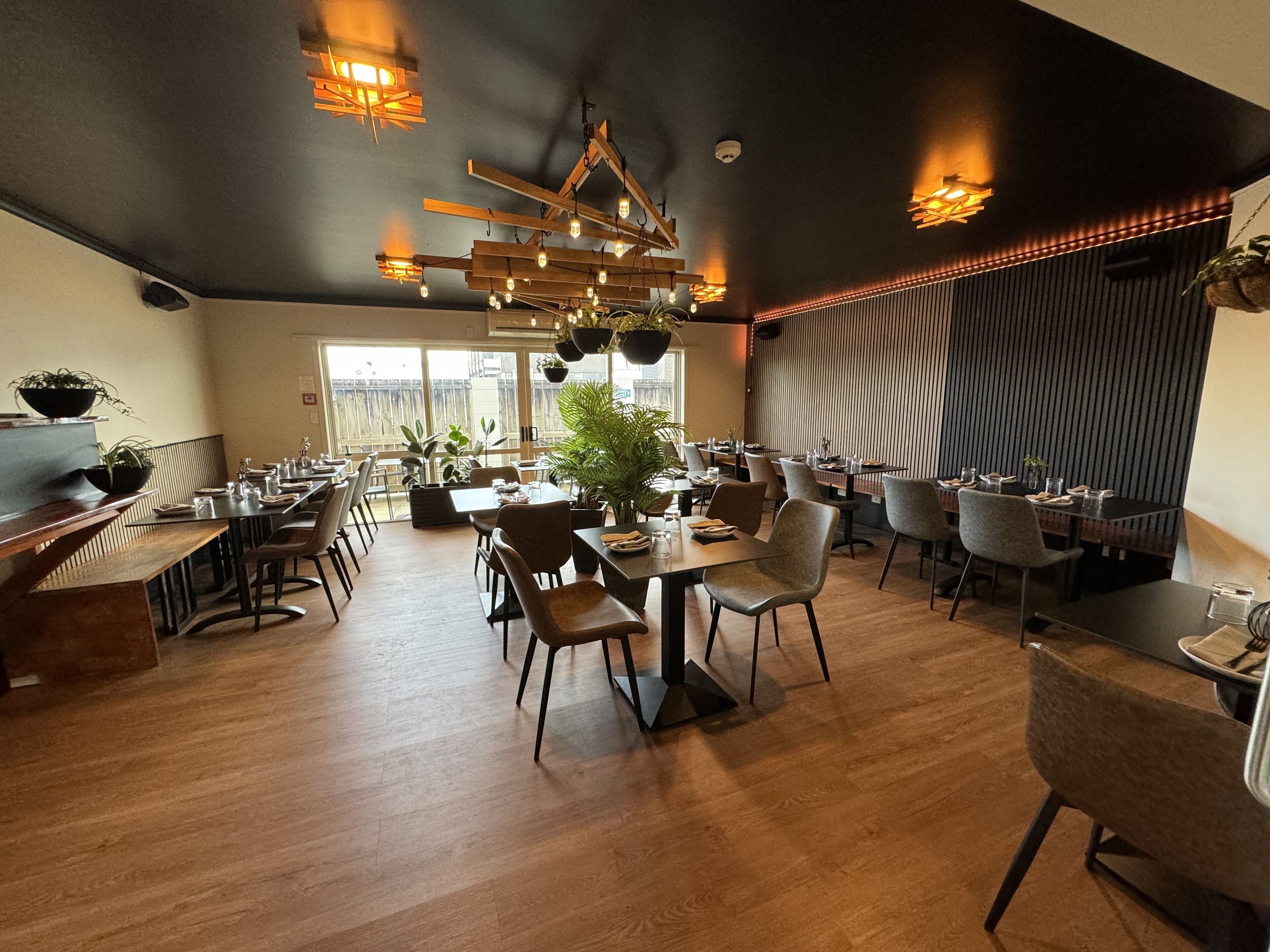 A spacious restaurant with wooden tables and chairs, potted plants, and modern lighting fixtures.