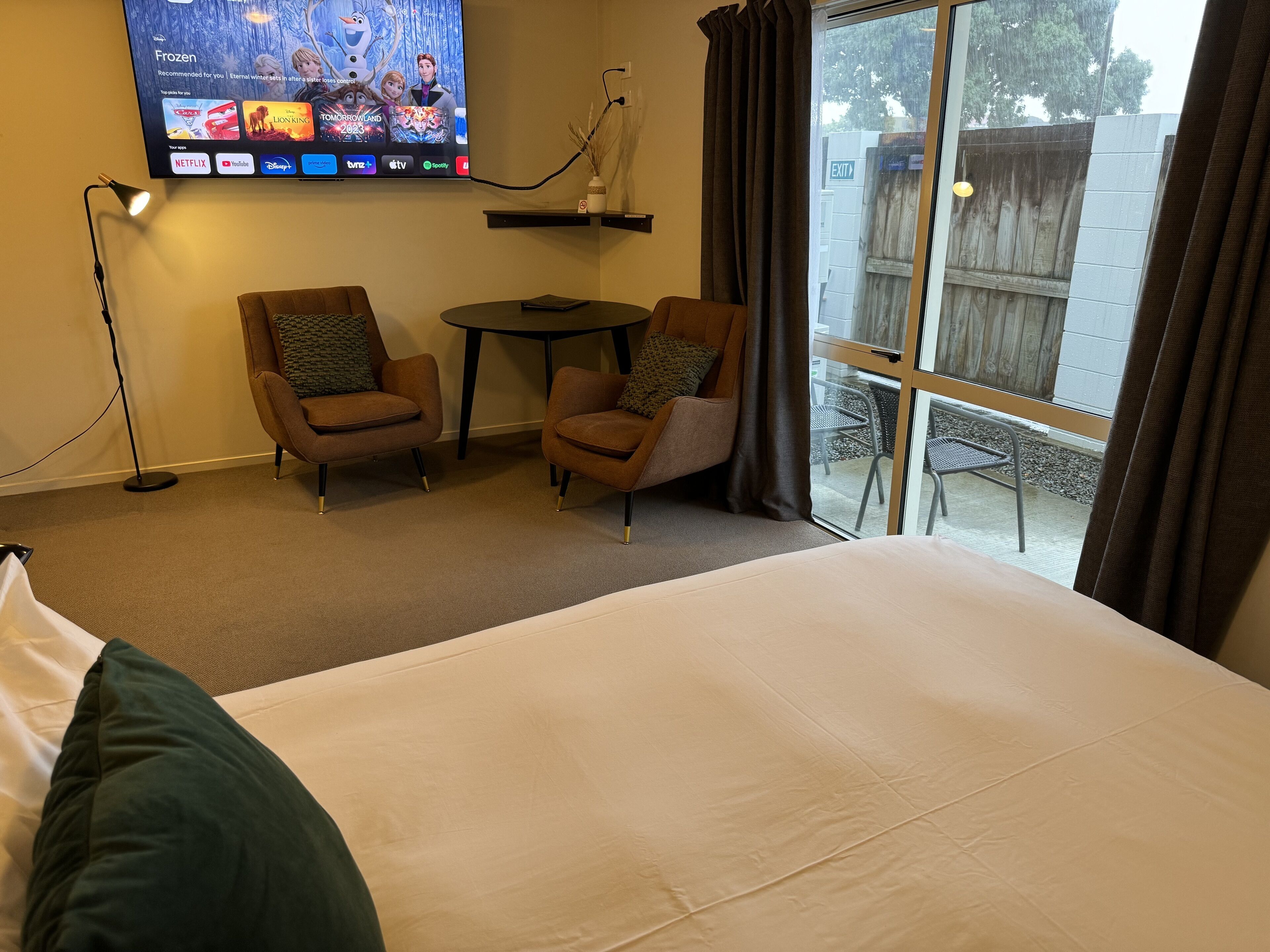 Habitación de hotel con cama, dos sillones, una mesita, televisión montada en la pared y una ventana con cortinas.