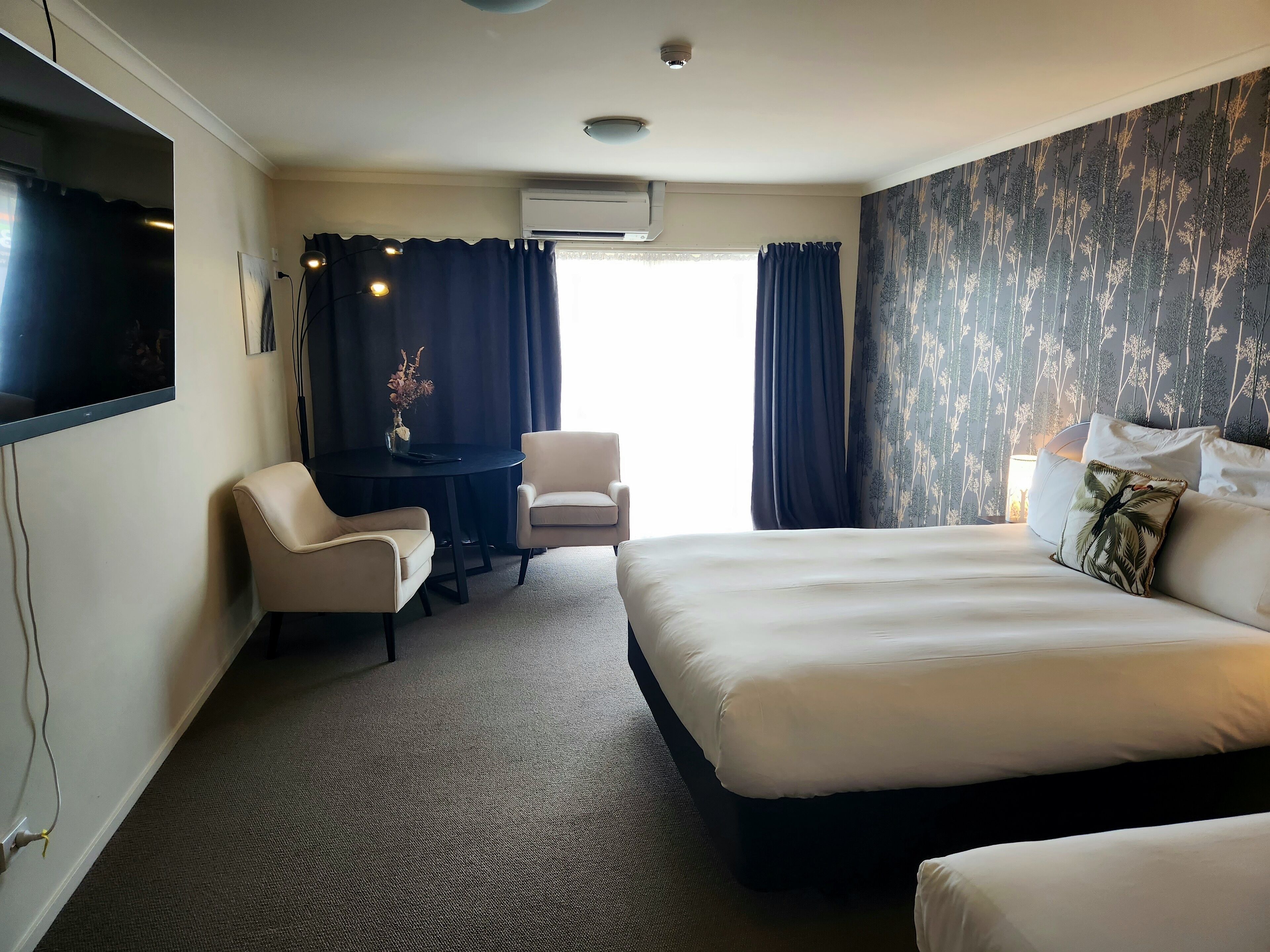 A hotel room with a bed, two armchairs, a small table with a vase, and a TV on the wall.