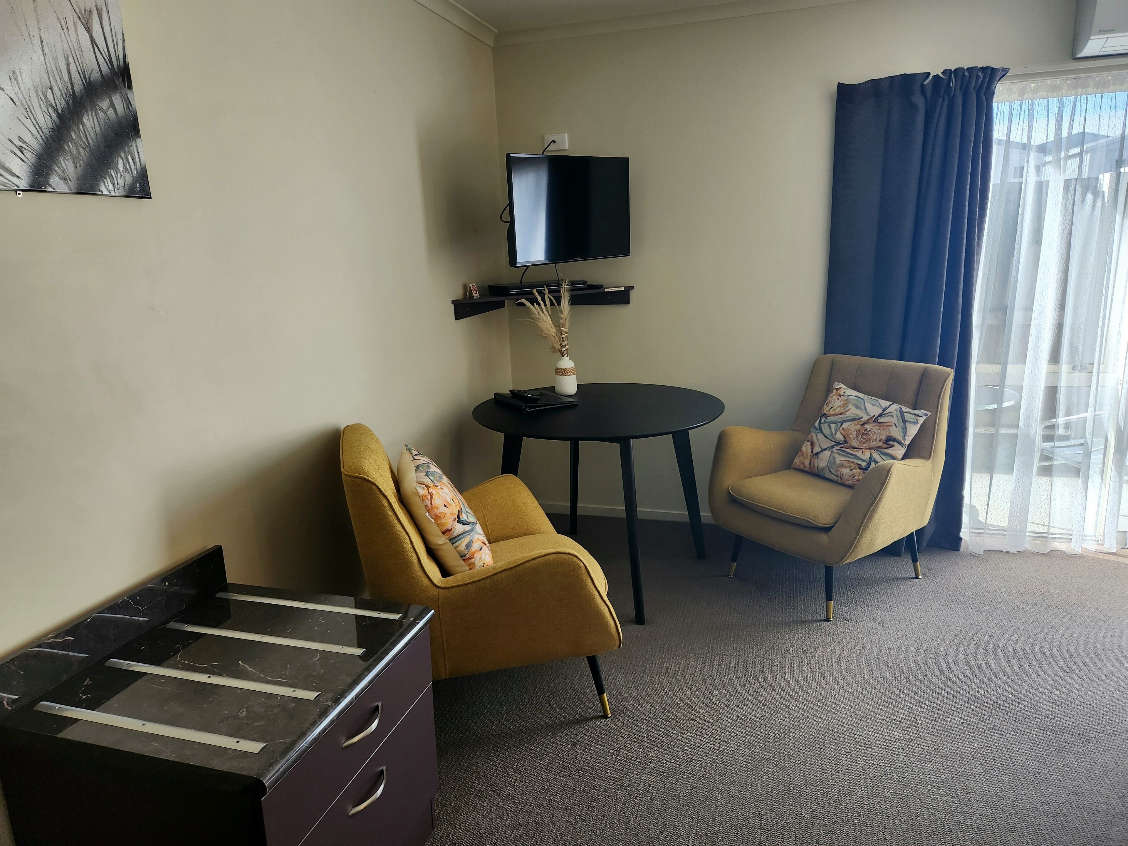 Et hotelværelse med sofa, stol, lille bord, fjernsyn og et vindue med gardiner.