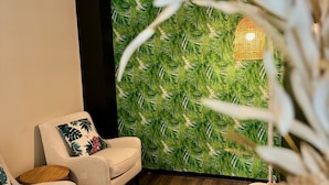A room with a wooden floor, a white armchair with a floral pillow, a wooden bucket, and a green leaf-patterned wall.