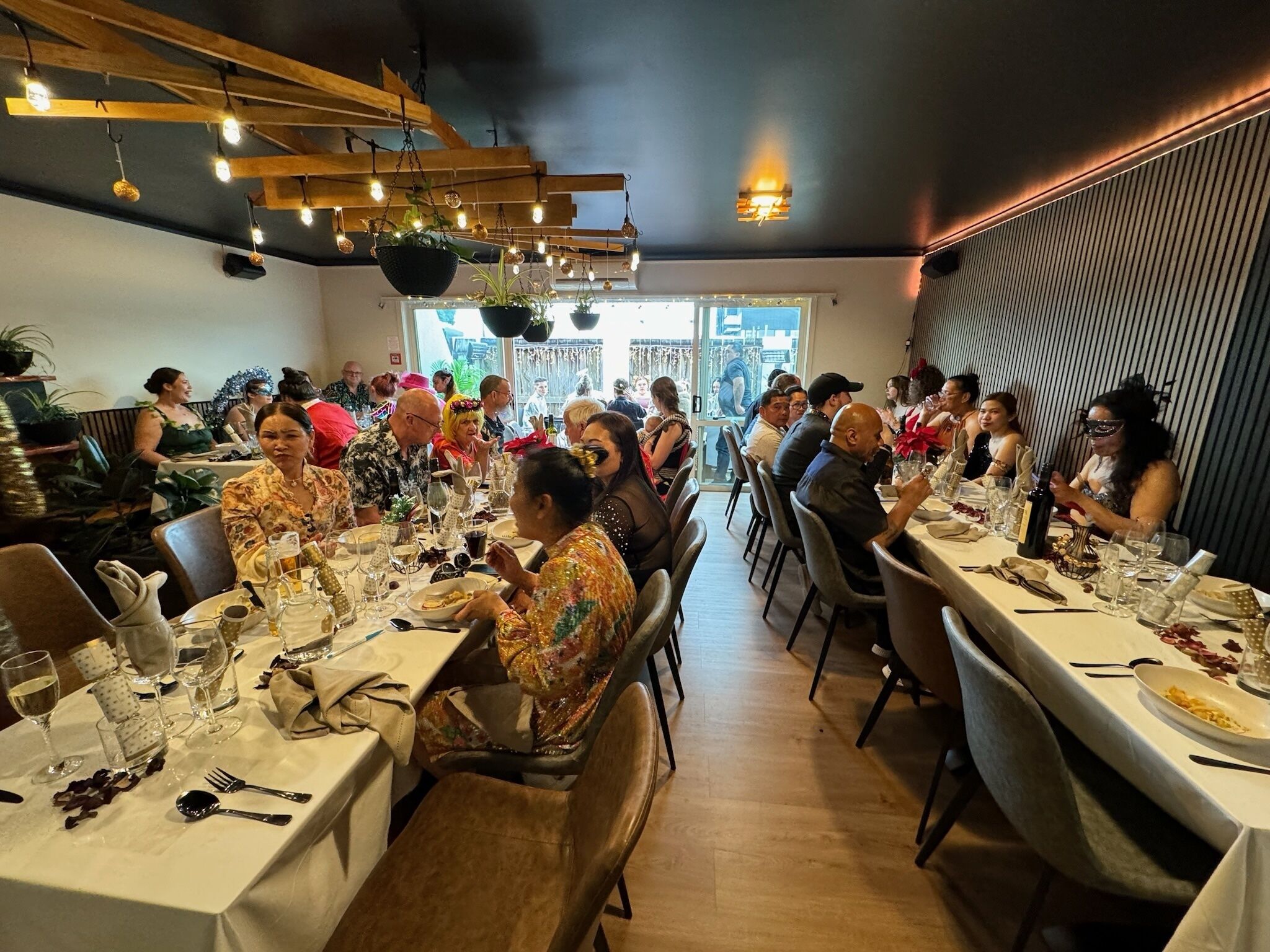 A restaurant with multiple tables set for dining, wooden ceiling beams, and hanging light fixtures.
