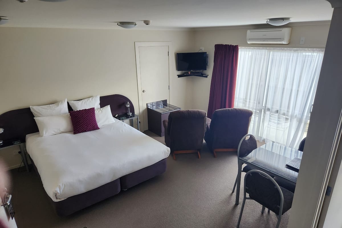 A hotel room with a bed, desk, chairs, a TV, and an air conditioning unit.