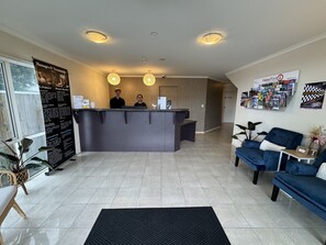 A reception area with a reception desk, two staff members, a bulletin board with information, a blue sofa, and a plant. - BKs Premier Motel Palmerston North (Palmerston North)