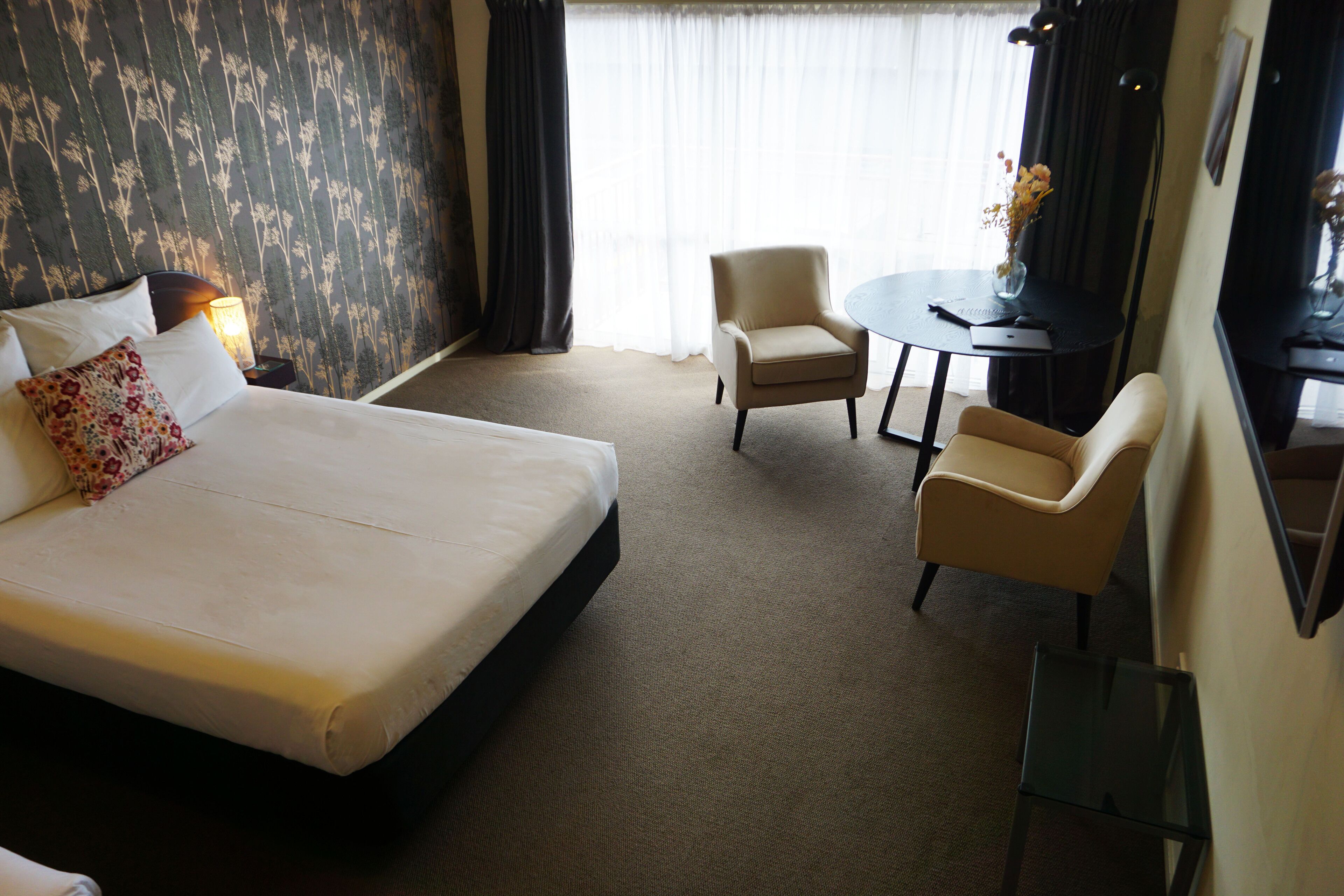 Quarto de hotel com uma cama grande, duas poltronas, uma mesa pequena e uma escrivaninha com uma luminária.